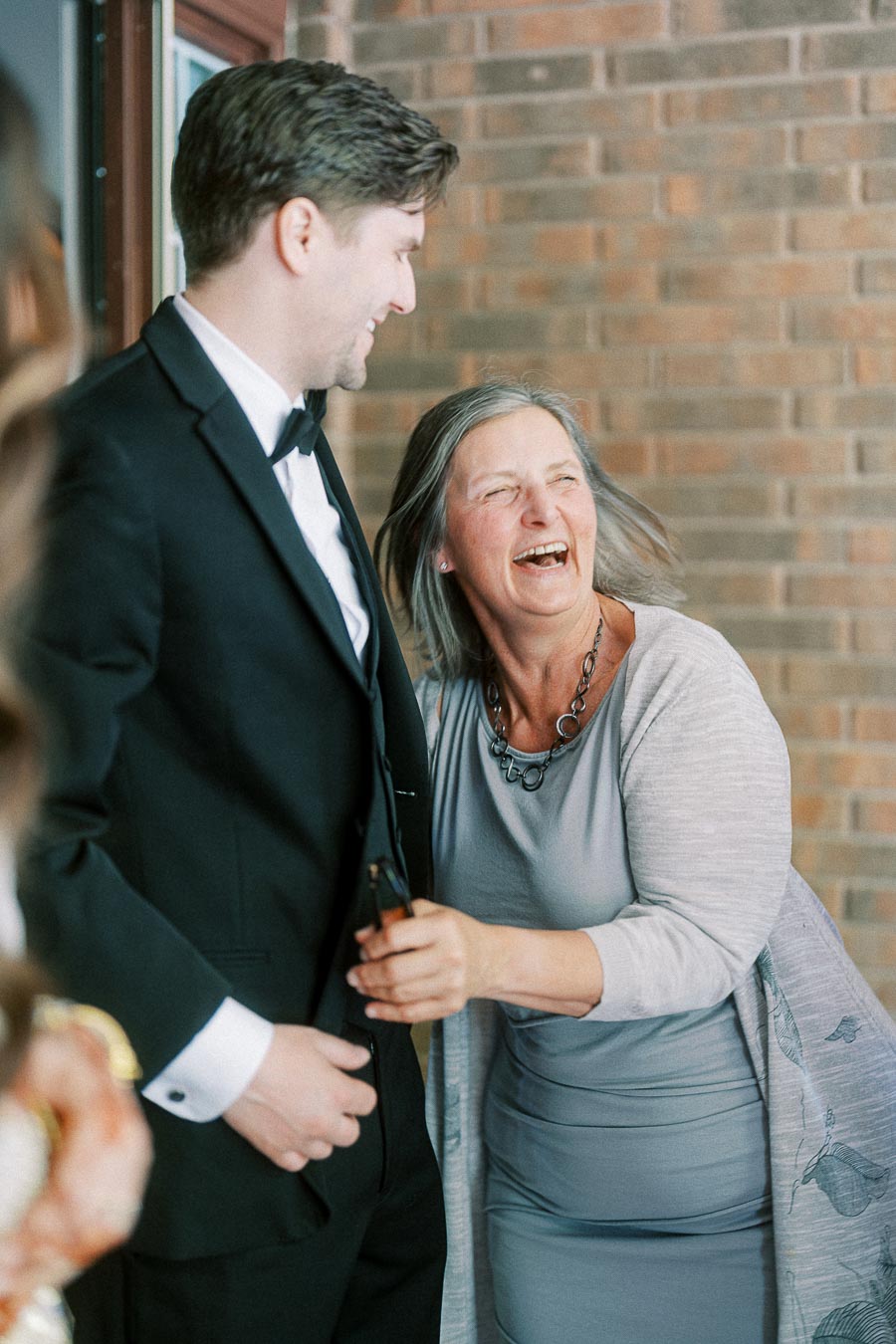 A joyful moment between two people at a formal event, featuring an elegantly dressed man in a suit and a laughing woman in a gray dress and cardigan, standing against a brick wall.