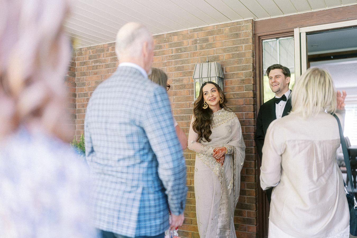 Elegant couple greeting guests at home, woman in traditional attire with henna designs, man in formal suit, creating a warm and welcoming atmosphere against a brick wall backdrop.