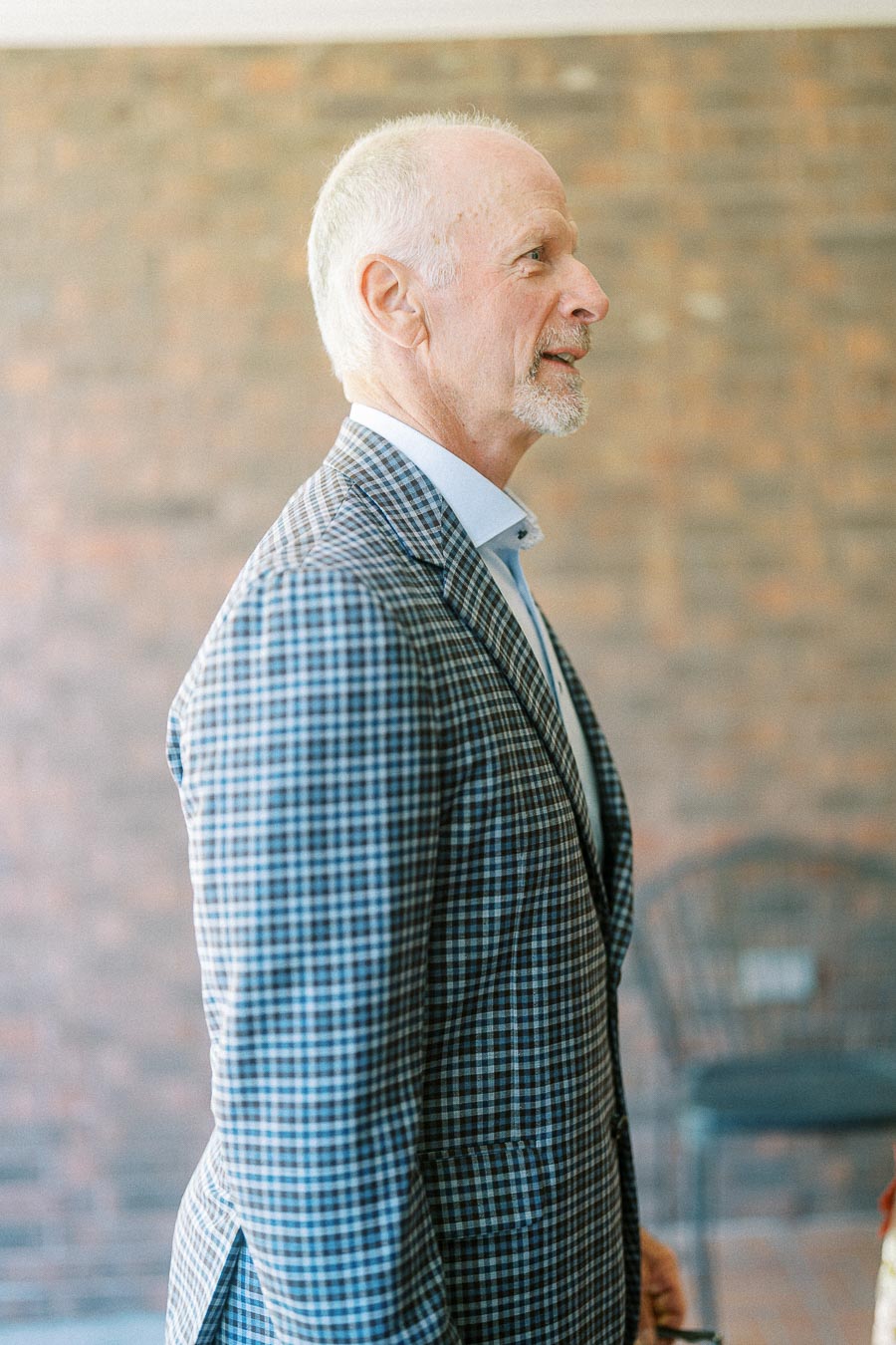 Elderly man in a stylish checkered suit standing indoors against a brick wall background.