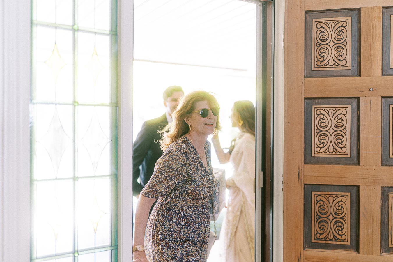 Woman in a floral dress and sunglasses entering a sunlight-lit doorway, with intricate wooden door design and stained glass window.