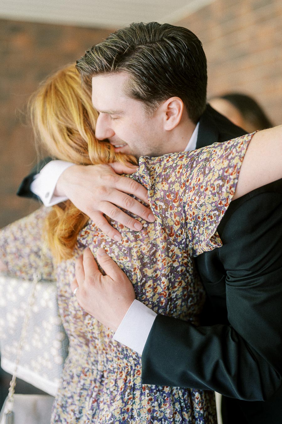 A man in a black suit warmly embraces a woman in a floral dress, capturing a heartfelt moment of affection and connection.