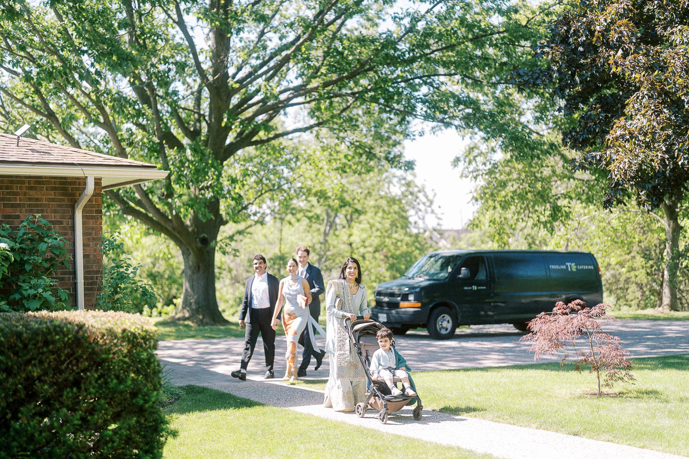A group of people enjoying a sunny day walk outside a brick house, with one woman pushing a stroller and others following her along a tree-lined street. A black van labeled Trellini Catering is parked nearby.