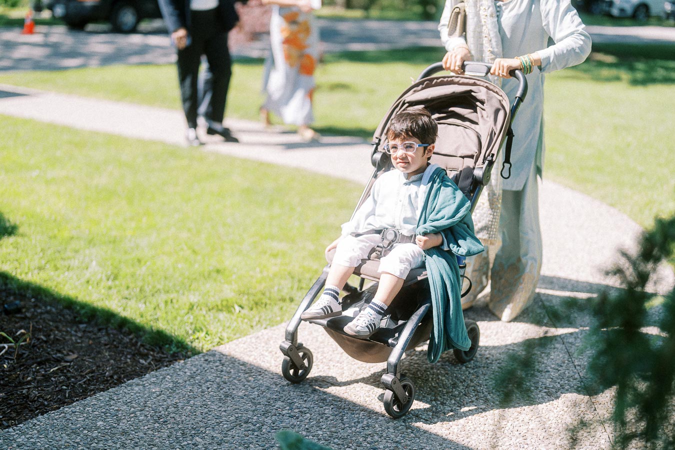 A young child wearing glasses sits comfortably in a stroller, dressed in light-colored clothes with a blue jacket draped over one shoulder, while a parent in traditional attire pushes the stroller along a sunny garden path.
