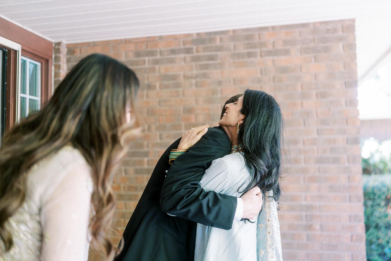 A heartfelt moment captured as two individuals embrace warmly on a porch with a brick wall backdrop. The scene reflects genuine emotion, while another person in the foreground observes the exchange, enhancing the intimate atmosphere.