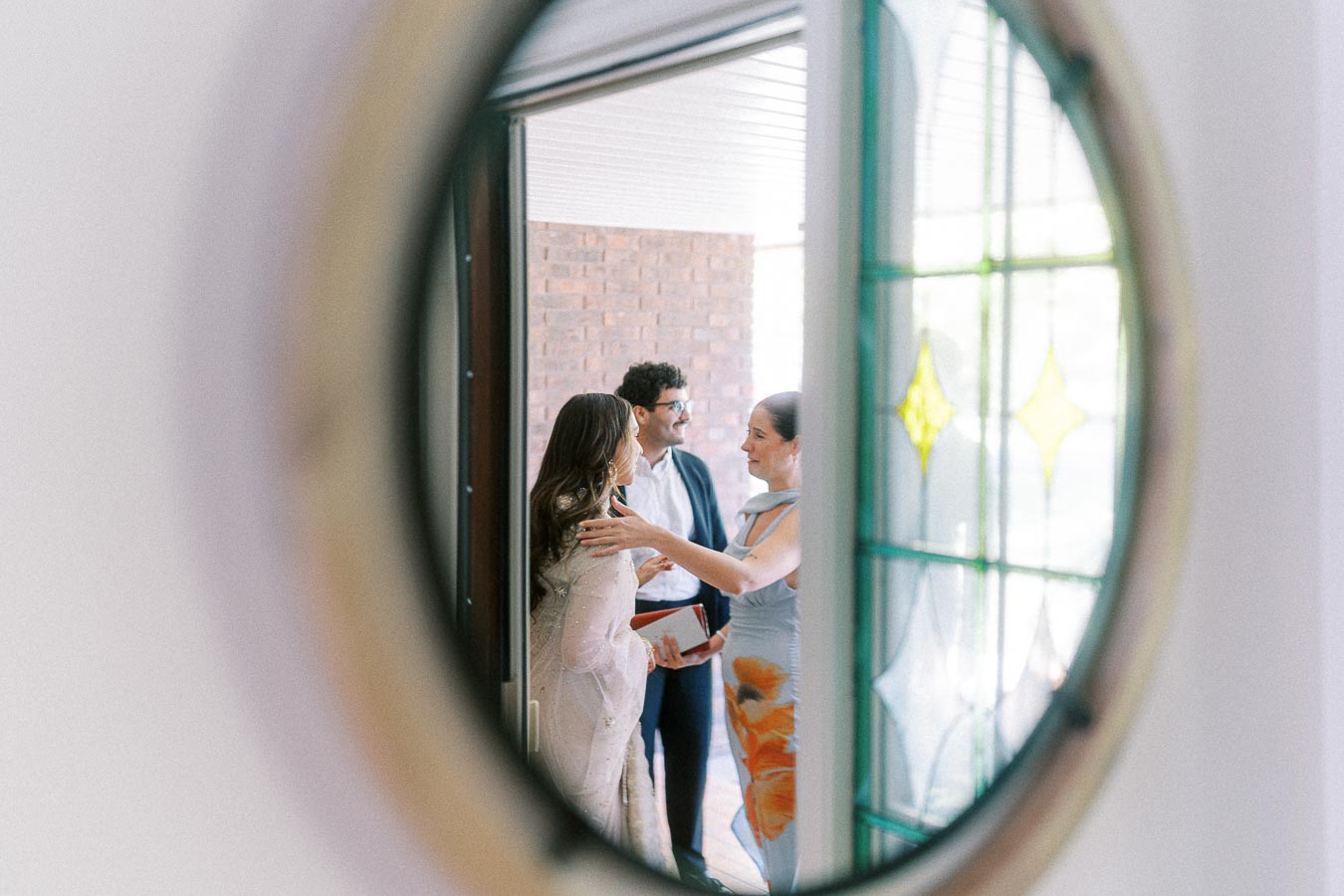 Three people are warmly greeting each other near an entrance, captured through a glass door with intricate stained patterns.