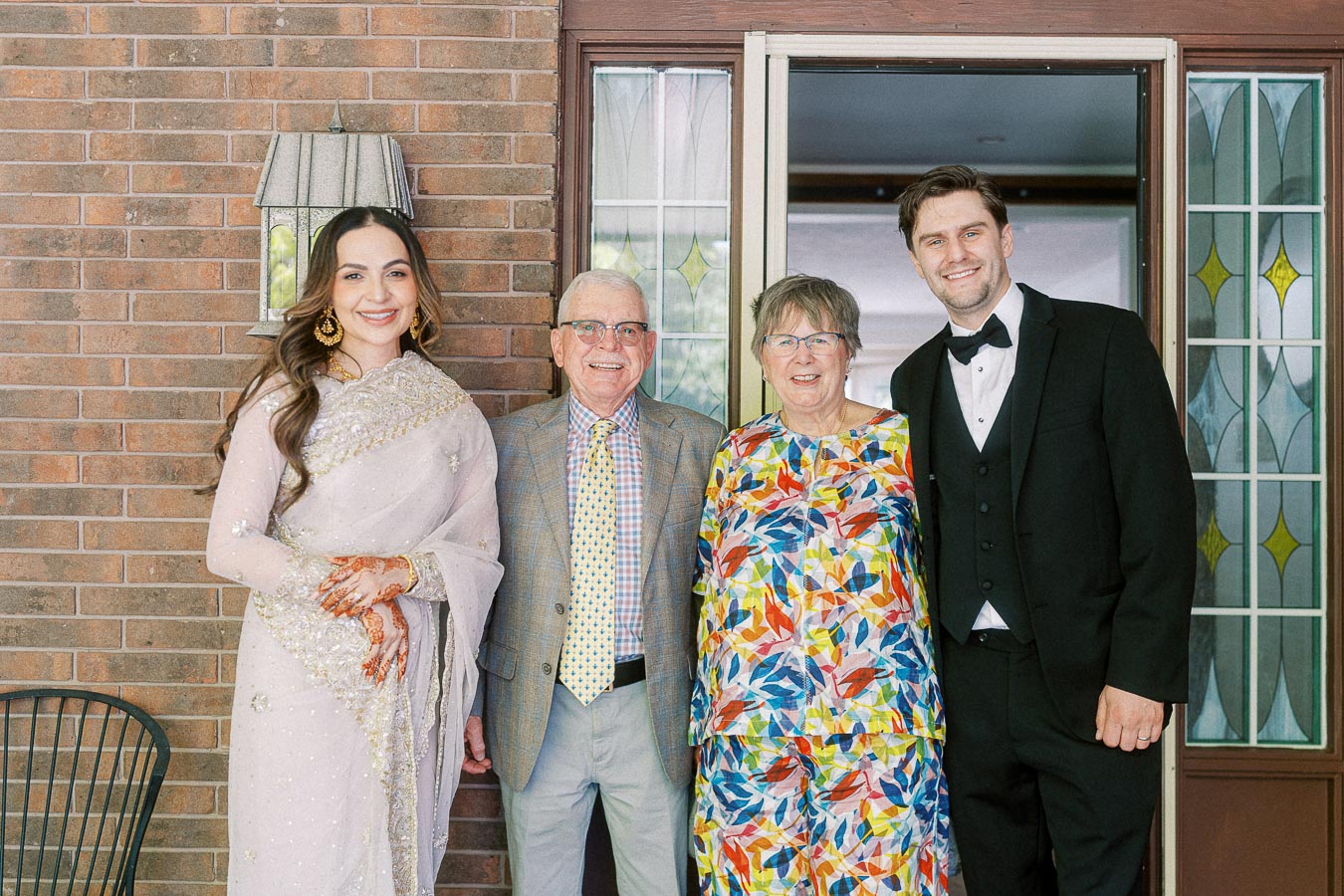 Elegant group at family event with diverse outfits including a traditional saree, colorful patterned dress, and formal tuxedo, posing happily in front of a brick wall and decorative glass door.