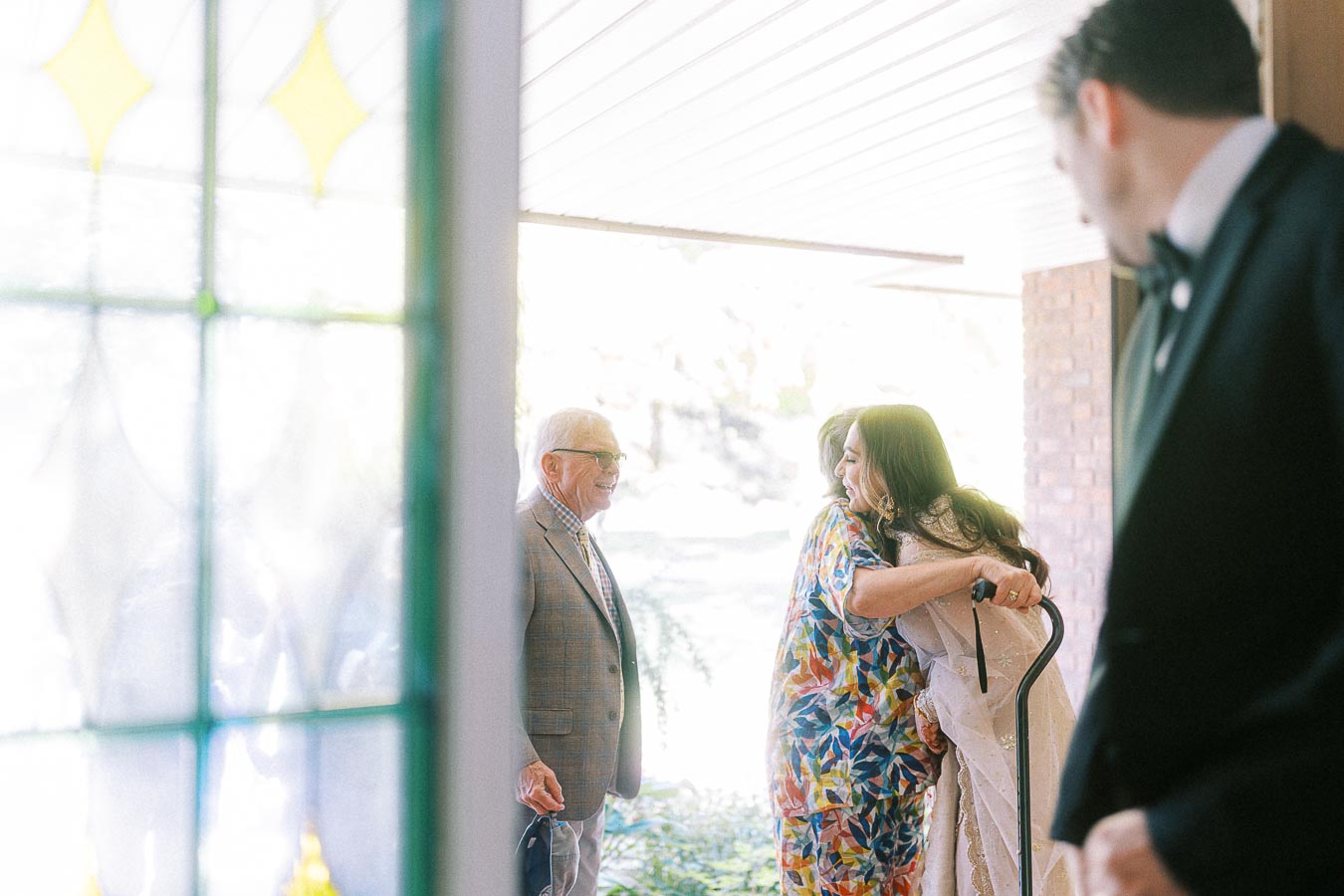 A warm family gathering at an entrance, with an elderly man in a plaid suit holding a walking stick, two women embracing, and a gentleman in a tuxedo looking on. Vibrant stained glass and soft lighting create an inviting atmosphere.