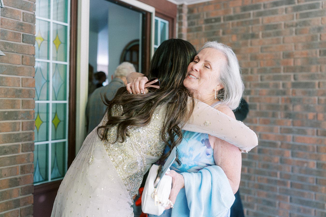 Two women embrace warmly outside a building, conveying a sense of friendship and happiness. One woman wears a detailed embroidered outfit, while an elderly woman in a blue dress smiles. The background features a brick wall and a stained glass door.