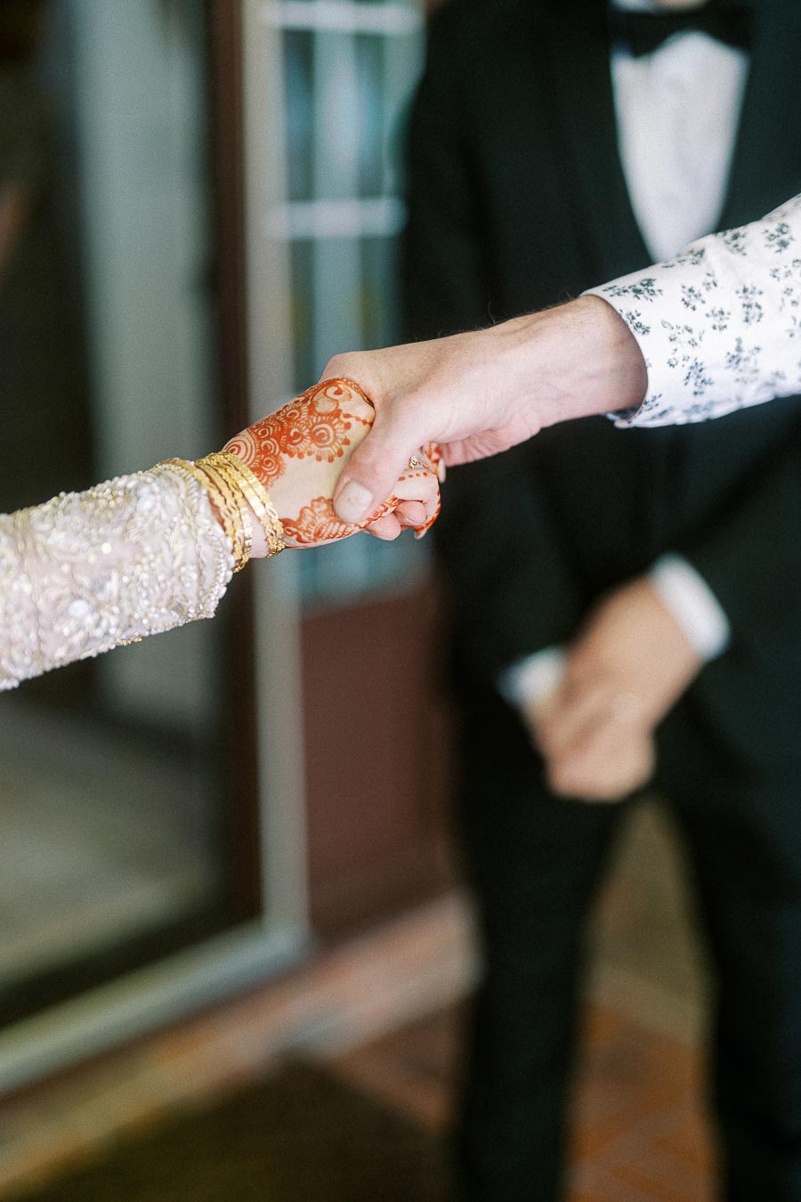 Close-up of a couple holding hands, with intricate henna designs and gold bangles on one hand, symbolizing cultural traditions and celebration.