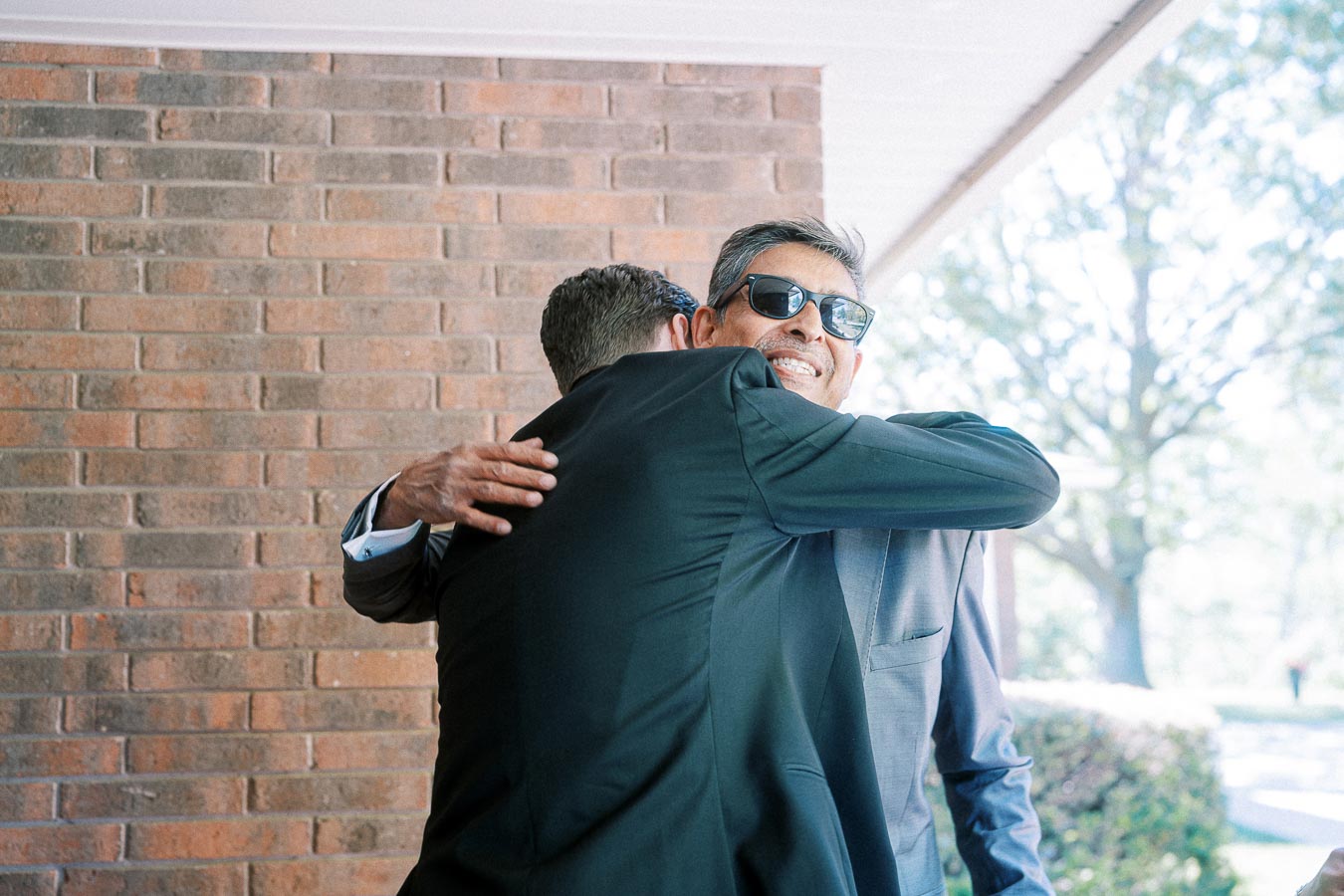 Two men in suits sharing a friendly hug outdoors next to a brick wall, with one wearing sunglasses and smiling warmly.