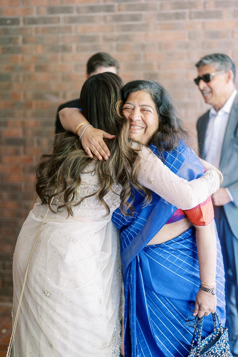 Two women embracing warmly, one in a white and gold traditional outfit and the other in a blue saree, with smiling expressions and a brick wall background.