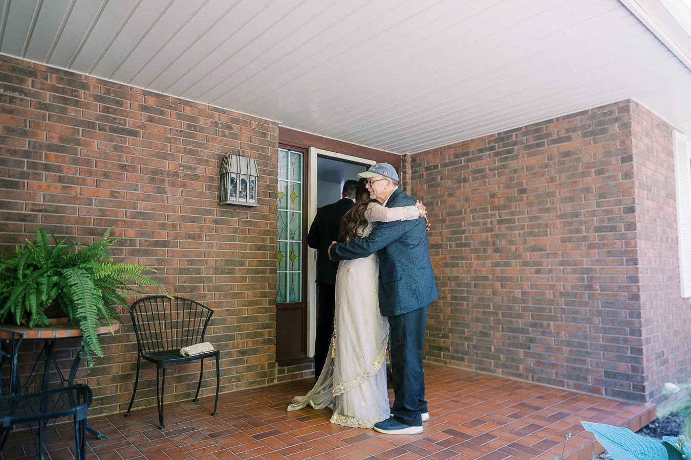 A bride and two men share an embrace on a brick porch near a decorative stained glass door, with potted plants and wrought iron chairs enhancing the cozy outdoor setting.