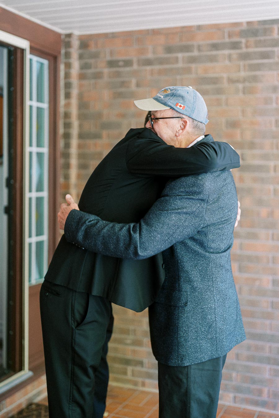 Two individuals warmly embrace in front of a brick wall, symbolizing friendship and connection. One person wears a casual hat, adding a touch of informality to the heartfelt moment.