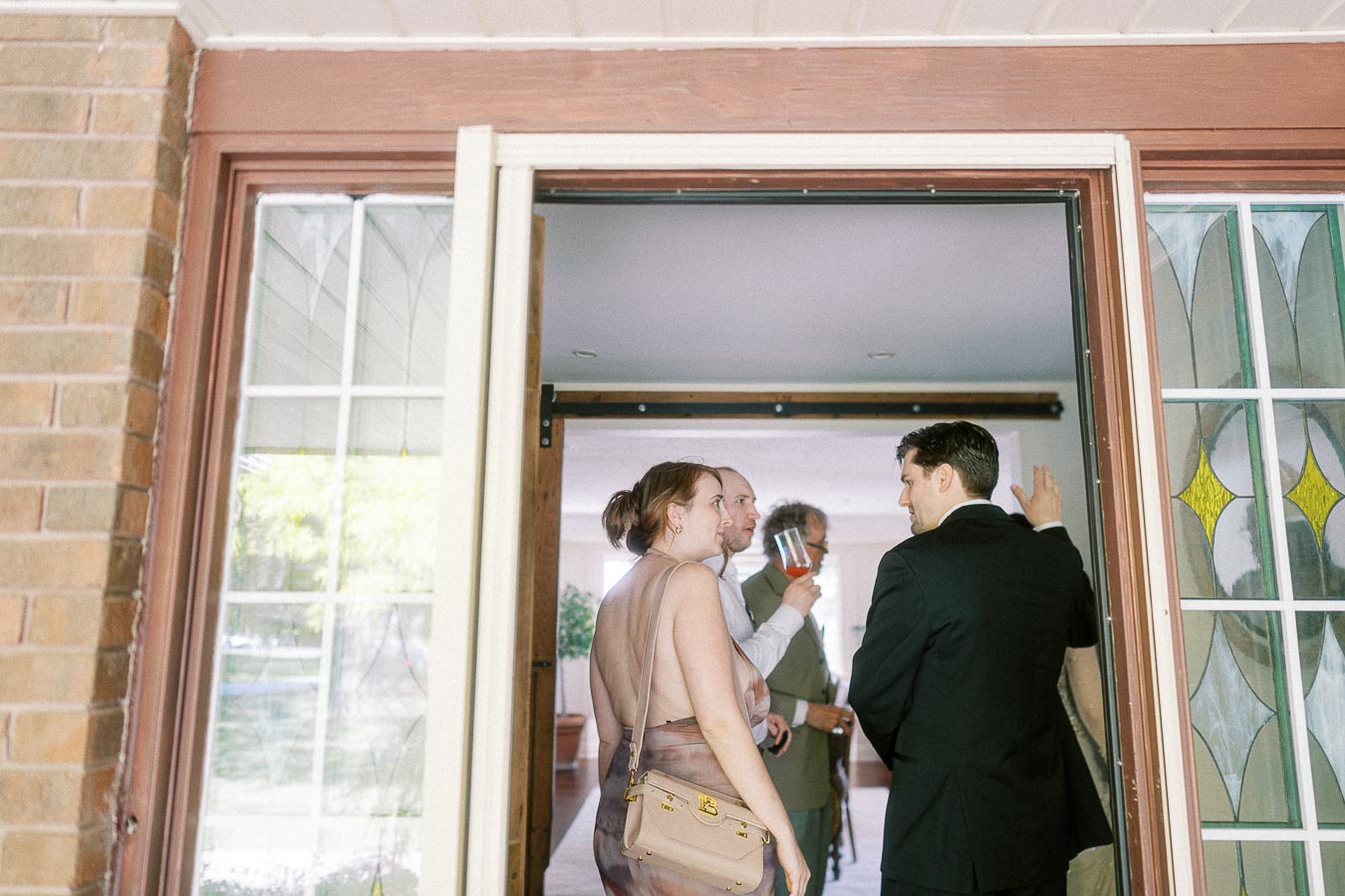 A group of people dressed in formal attire socializing near an open doorway, with sunlight streaming through stained glass windows.