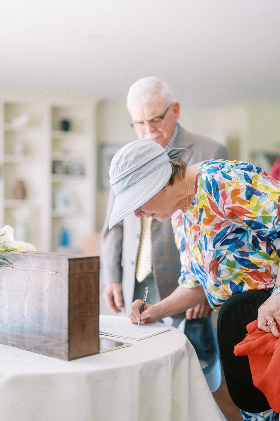 Elderly woman in colorful shirt signing a document on a table, with an elderly man in a suit watching in a bright, modern room.
