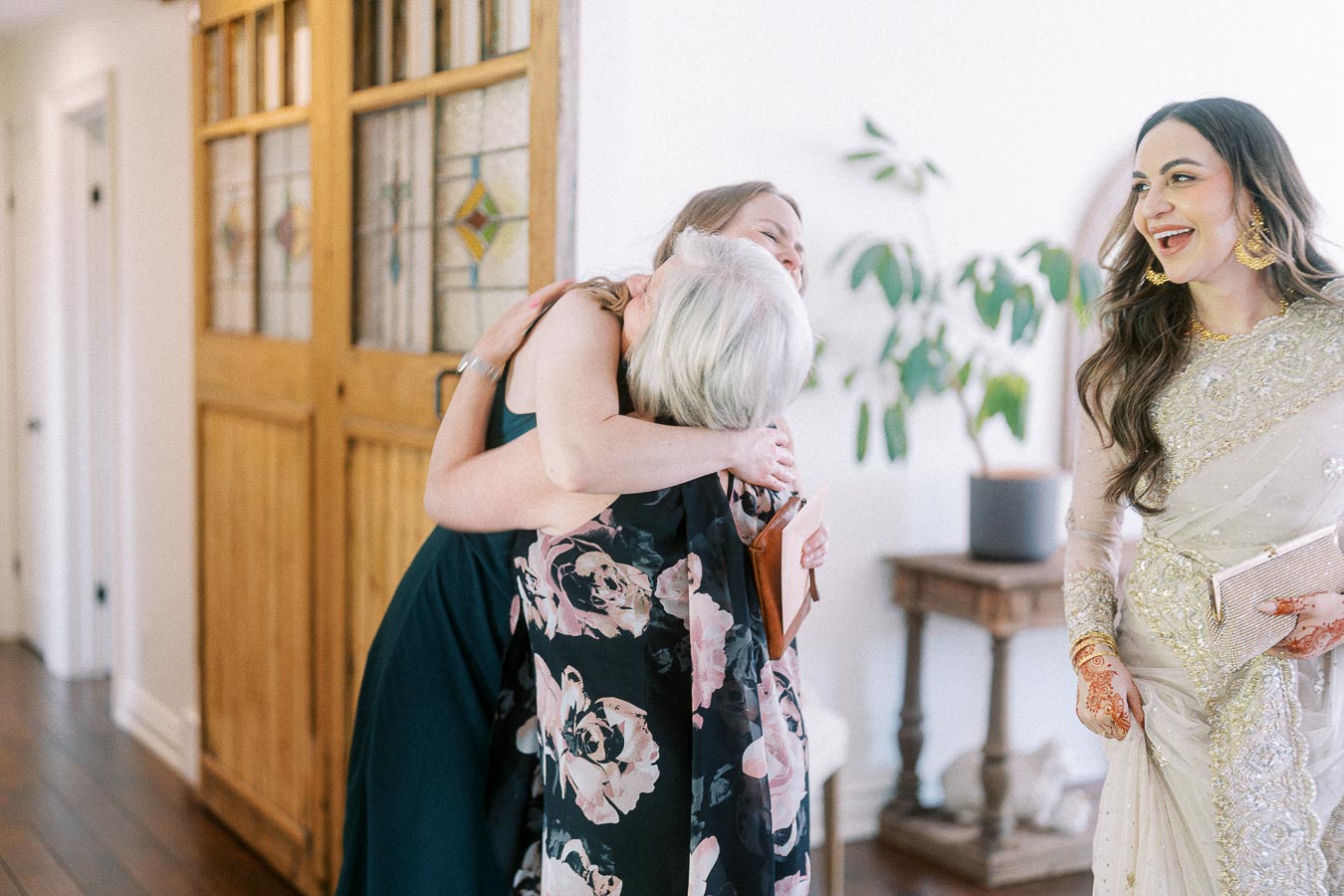 Three women share a joyful moment in an elegantly decorated room. One, in a floral dress, embraces another wearing a dark dress, while a smiling woman in a white and gold embroidered outfit looks on. The atmosphere is filled with warmth and happiness, complemented by the background of stained glass doors and potted plants.