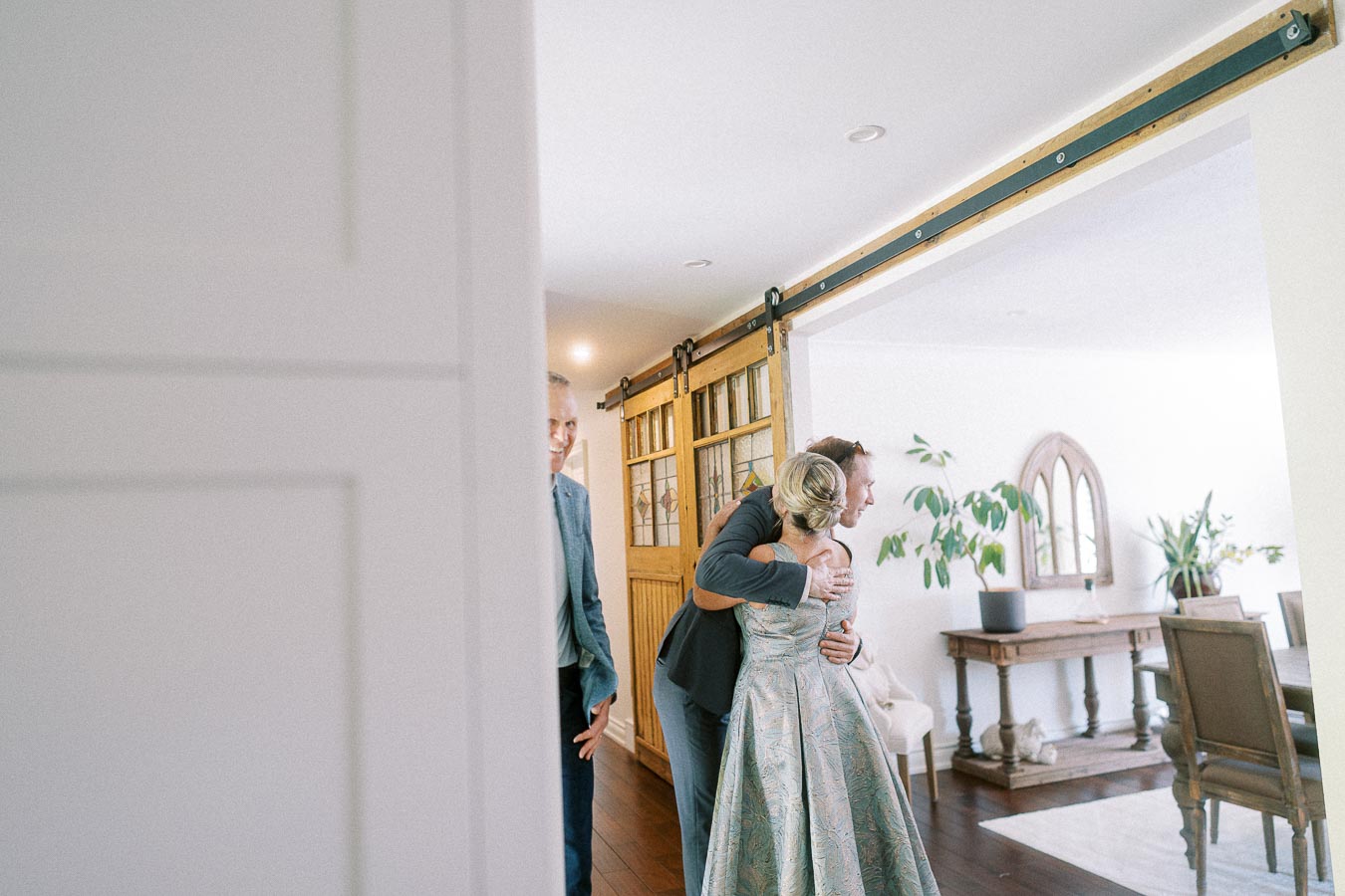 A person in a formal suit warmly embraces a woman in an elegant dress inside a modern, stylishly decorated room featuring wooden floors and potted plants. A decorative wooden sliding door is positioned centrally, while another person in a suit appears smiling in the background.
