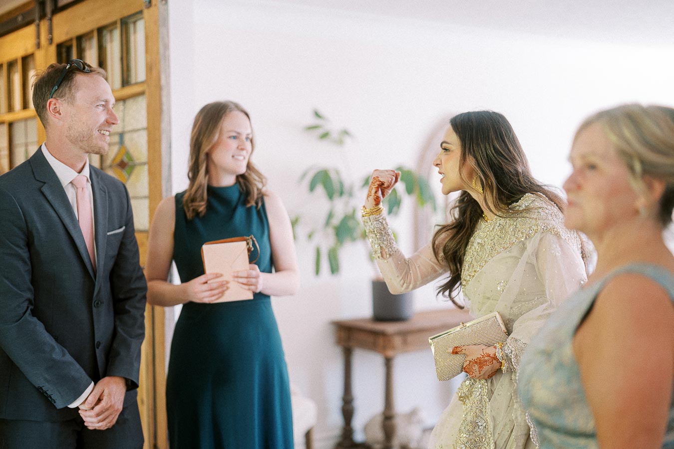 Group of people at an indoor gathering, featuring a woman in an elegant outfit with intricate embroidery and henna designs, talking animatedly while holding a clutch; others dressed formally, smiling, and conversing.