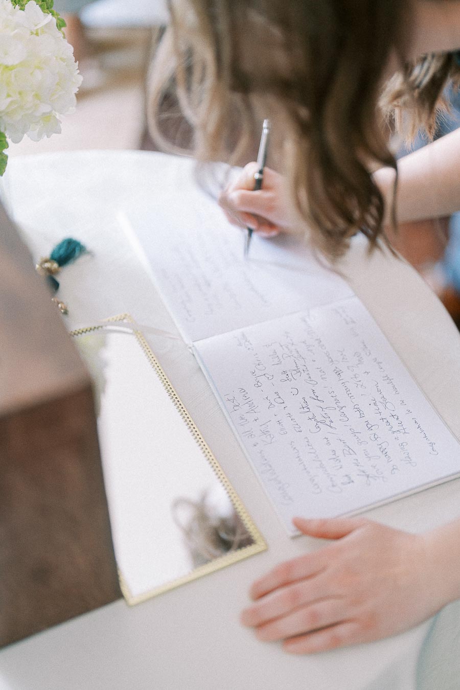 A person writing in a guestbook at an event, with a pen in hand and a mirror reflecting part of the scene, set on a white table accompanied by white flowers.