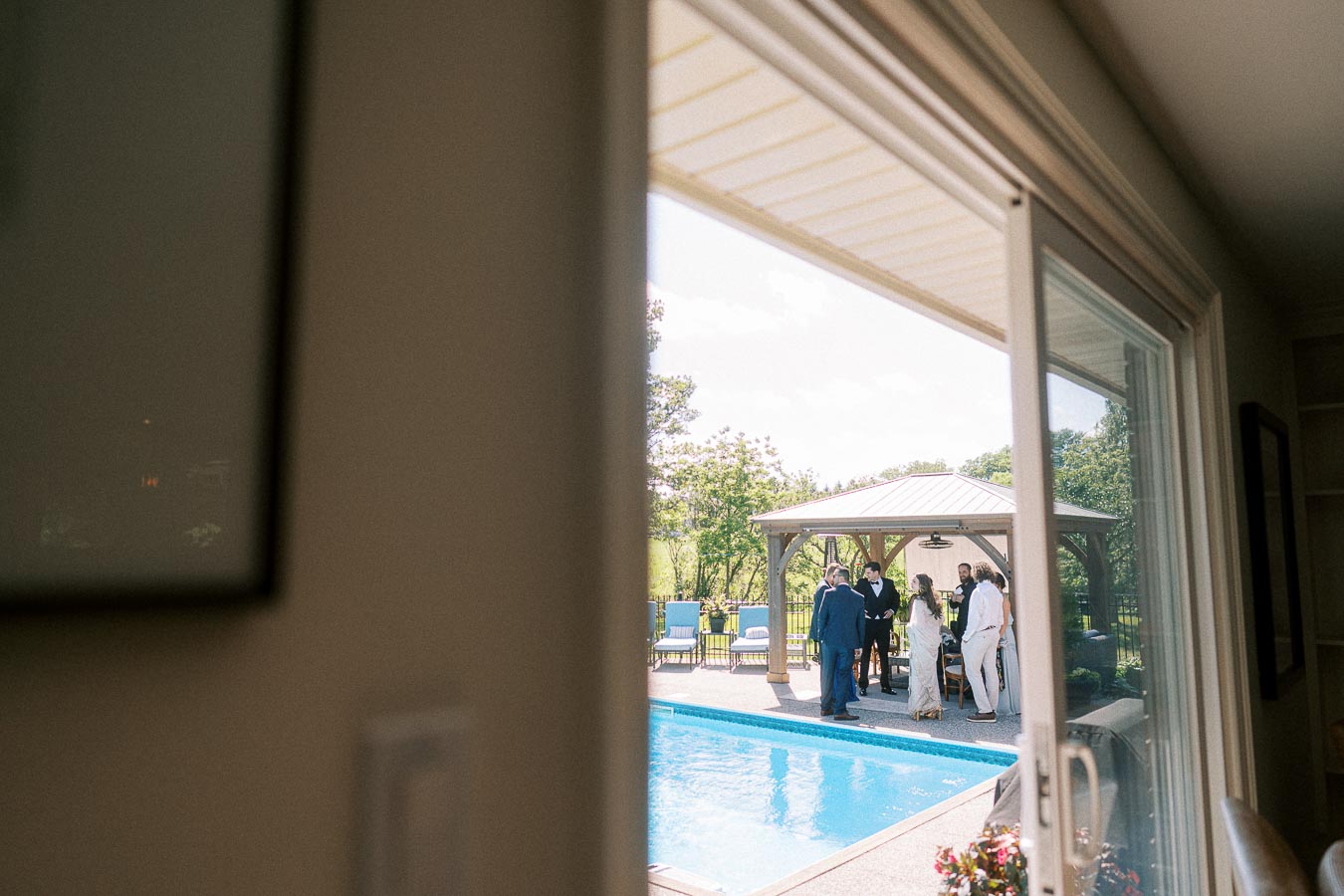 View from indoors through a sliding glass door, capturing an outdoor gathering by a swimming pool. People are standing under a gazebo, surrounded by greenery and outdoor furniture, on a sunny day.