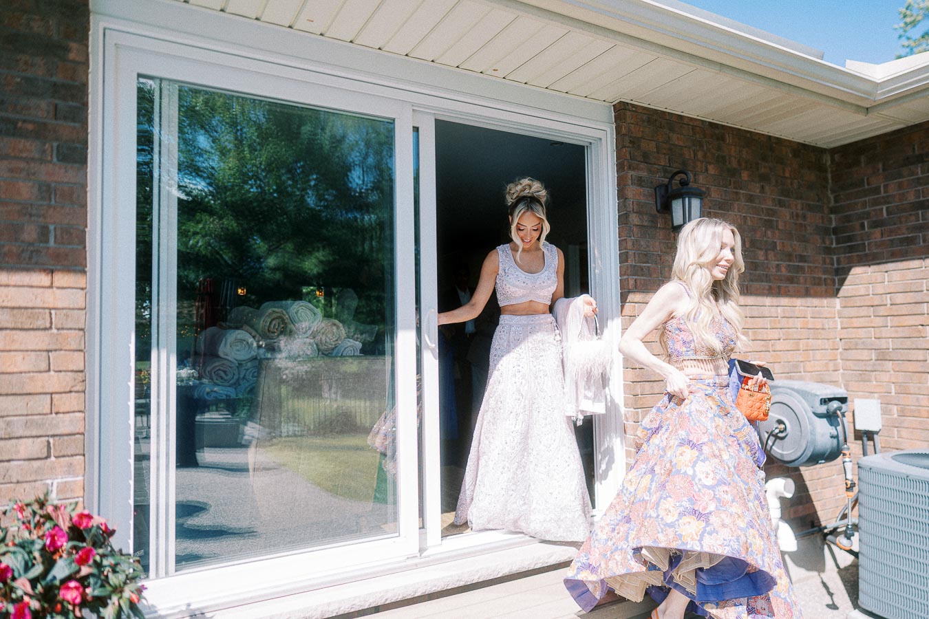 Two women in elegant floral dresses step out of a brick house on a sunny day, showcasing fashion and style.