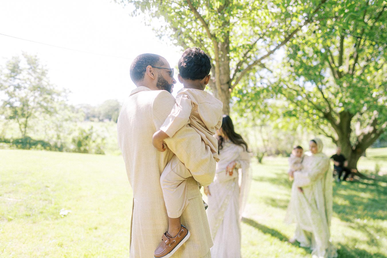 A man holding a child in a sunlit park, surrounded by trees, with other people in the background wearing traditional attire, highlighting a family gathering or outdoor celebration.