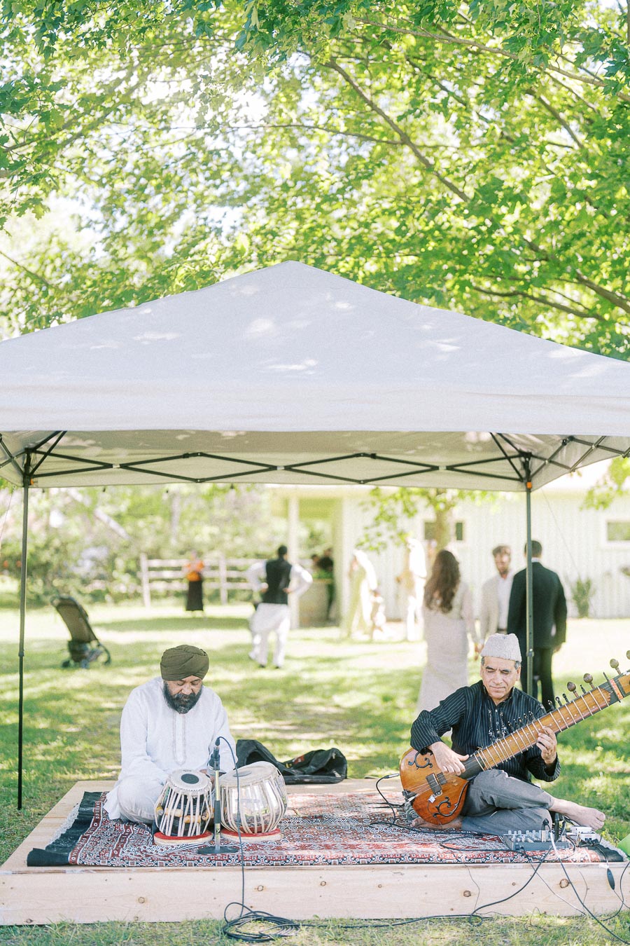 Outdoor event with musicians playing traditional Indian instruments under a white canopy, surrounded by greenery and attendees in the background.