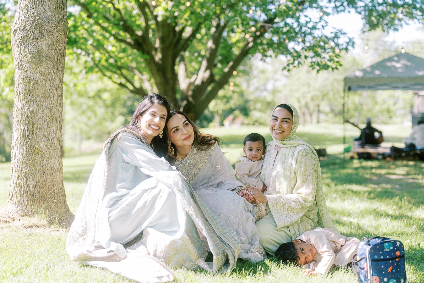 A joyful family gathering in a sunny park setting. Two women dressed in elegant traditional attire and two young children are seated on the grass, surrounded by trees. A picnic tent is visible in the background.