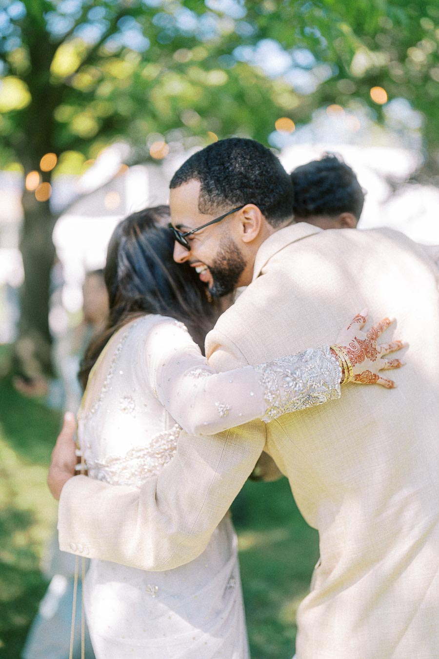 A joyful embrace between a man in a light beige suit and a woman wearing a glittering, embroidered dress with henna on her hands, captured in a sunlit garden setting, highlighting friendship and celebration.