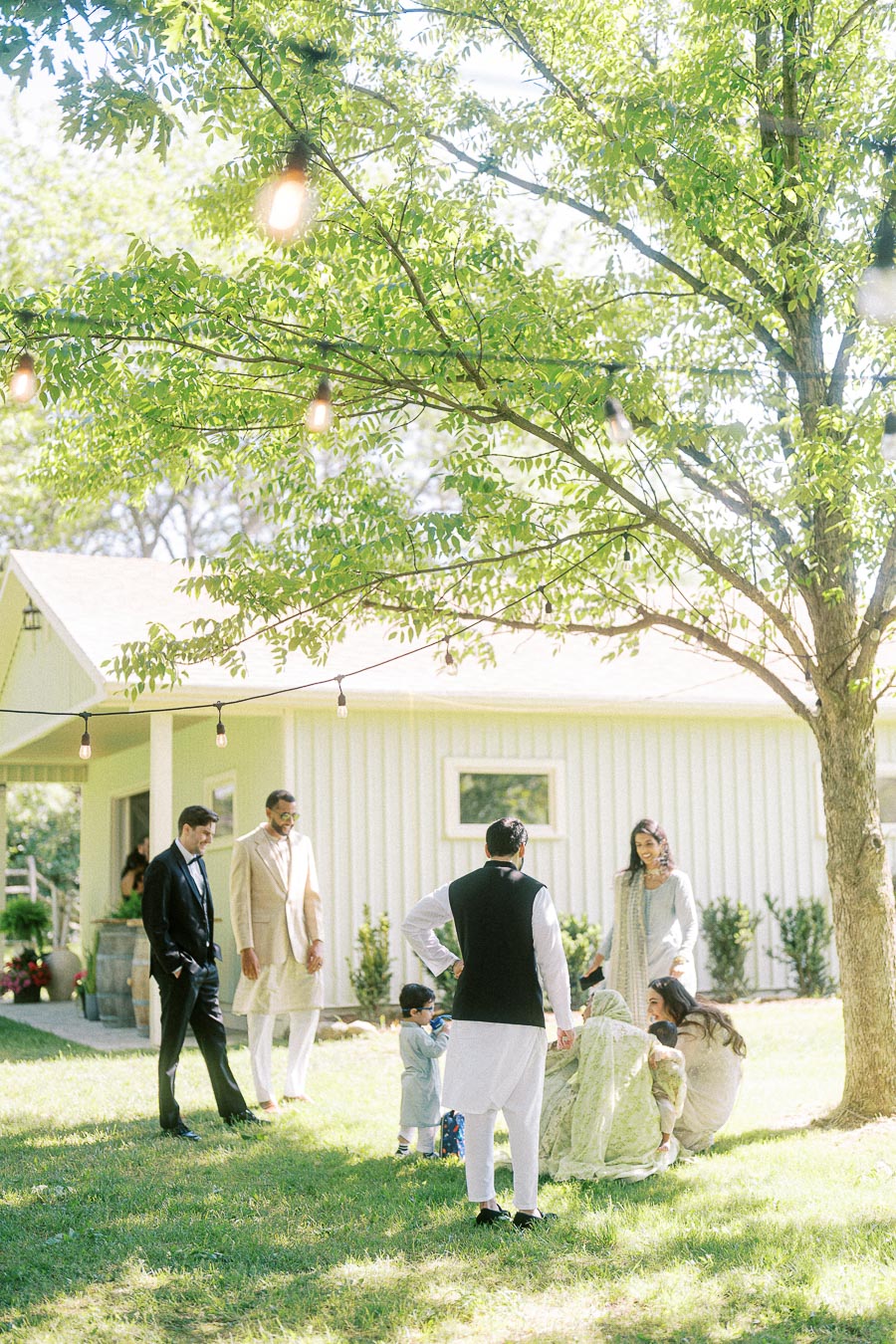 Group of people in traditional attire having a conversation outdoors in a garden setting with festive string lights and a white house in the background.