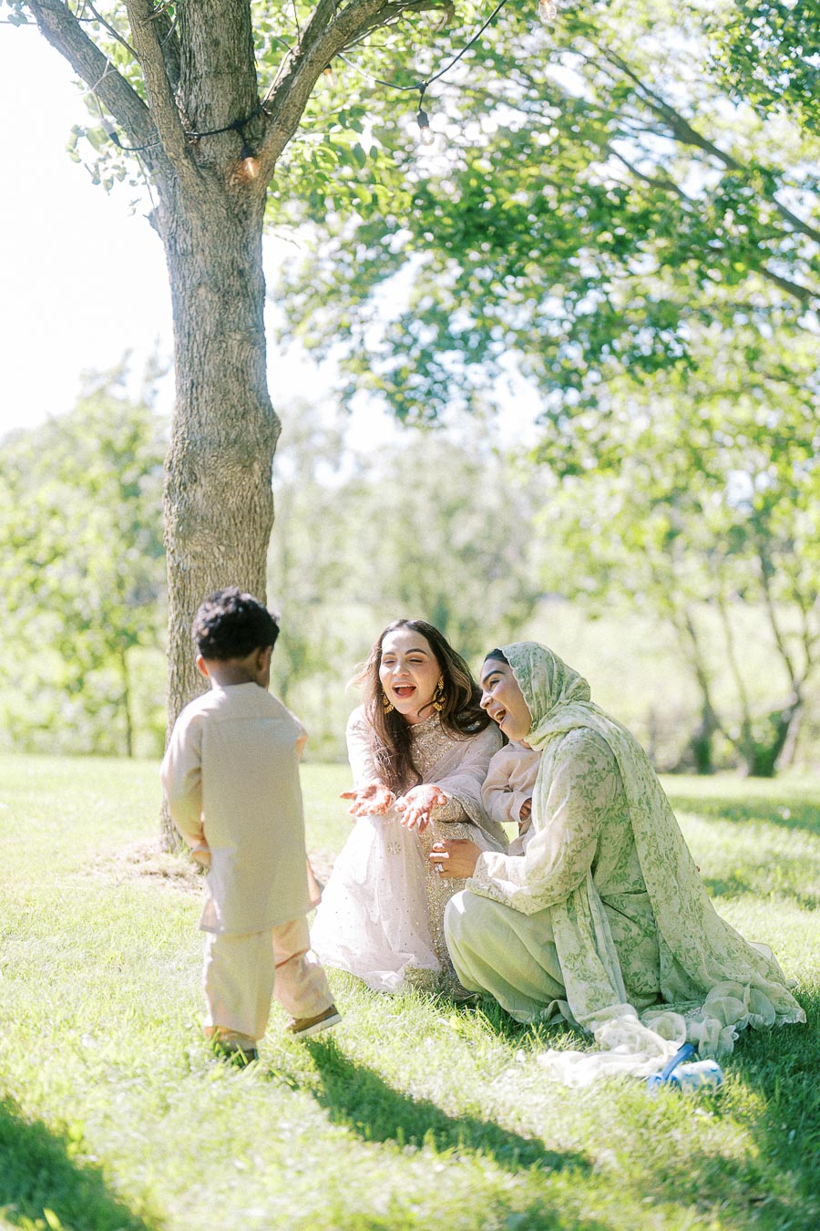 A joyful outdoor scene of two women wearing traditional clothing, smiling and engaging with a young child under a tree on a sunny day, highlighting a moment of family bonding and cultural celebration in a lush green park.