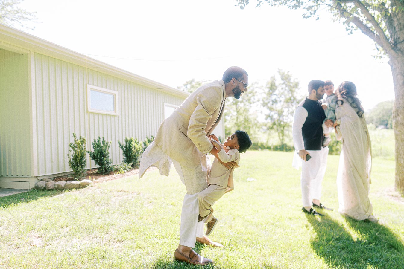 A joyful outdoor family gathering with adults and children dressed in elegant attire, enjoying playful moments on a sunny day near a metal building surrounded by greenery.