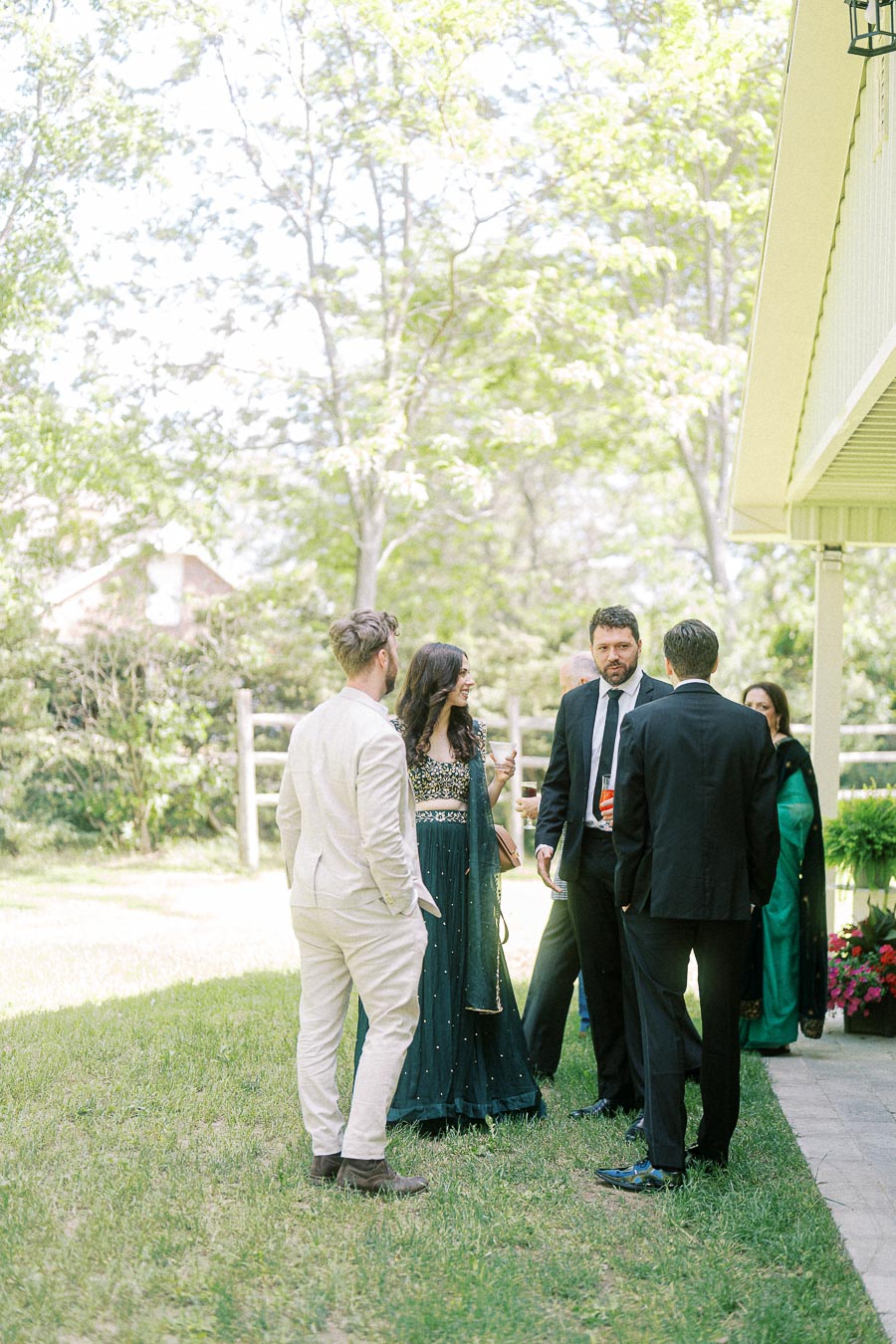Group of people socializing at an outdoor event, wearing formal and elegant attire, standing on a grassy area beside a house under a sunny sky.