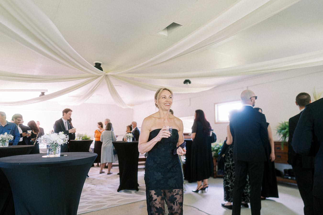 A woman in an elegant black dress holding a drink, smiling at an indoor formal gathering with guests mingling in the background.