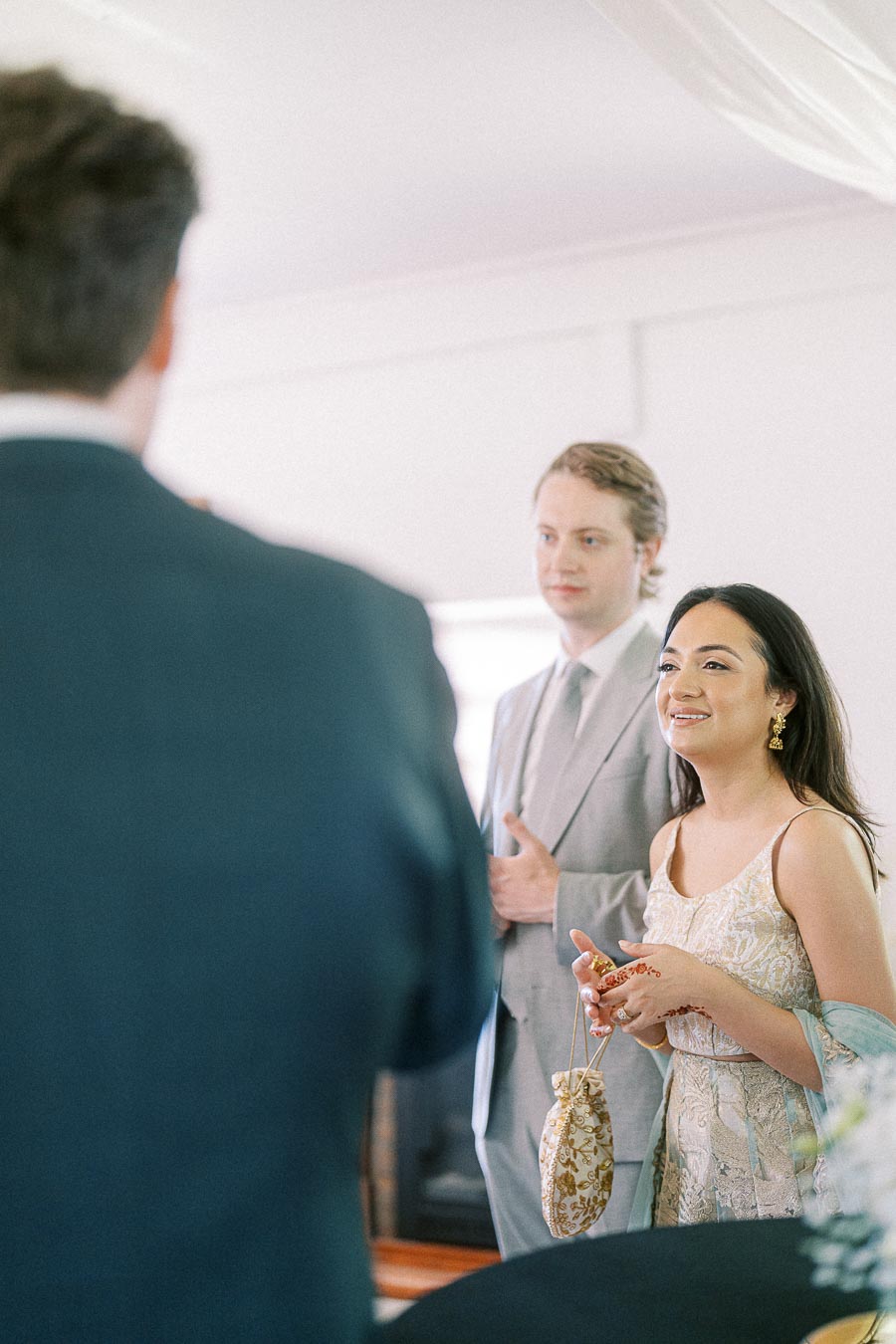 Guests at wedding reception, elegantly dressed woman holding a decorative pouch and smiling, accompanied by individual in a light gray suit, standing indoors with draped ceiling.