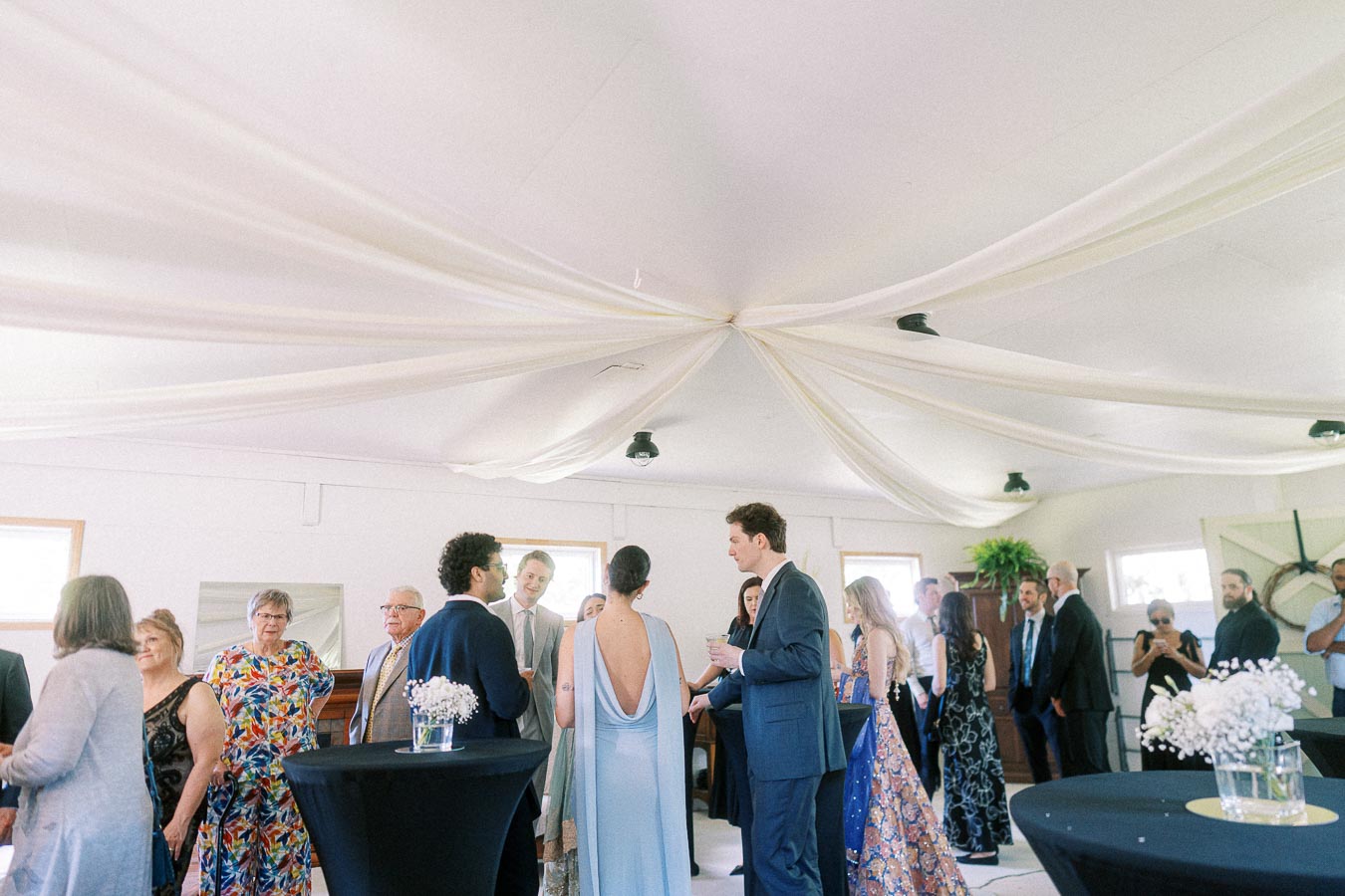 People mingling at an elegant indoor event with white draped ceiling decorations and round tables, dressed in formal attire.
