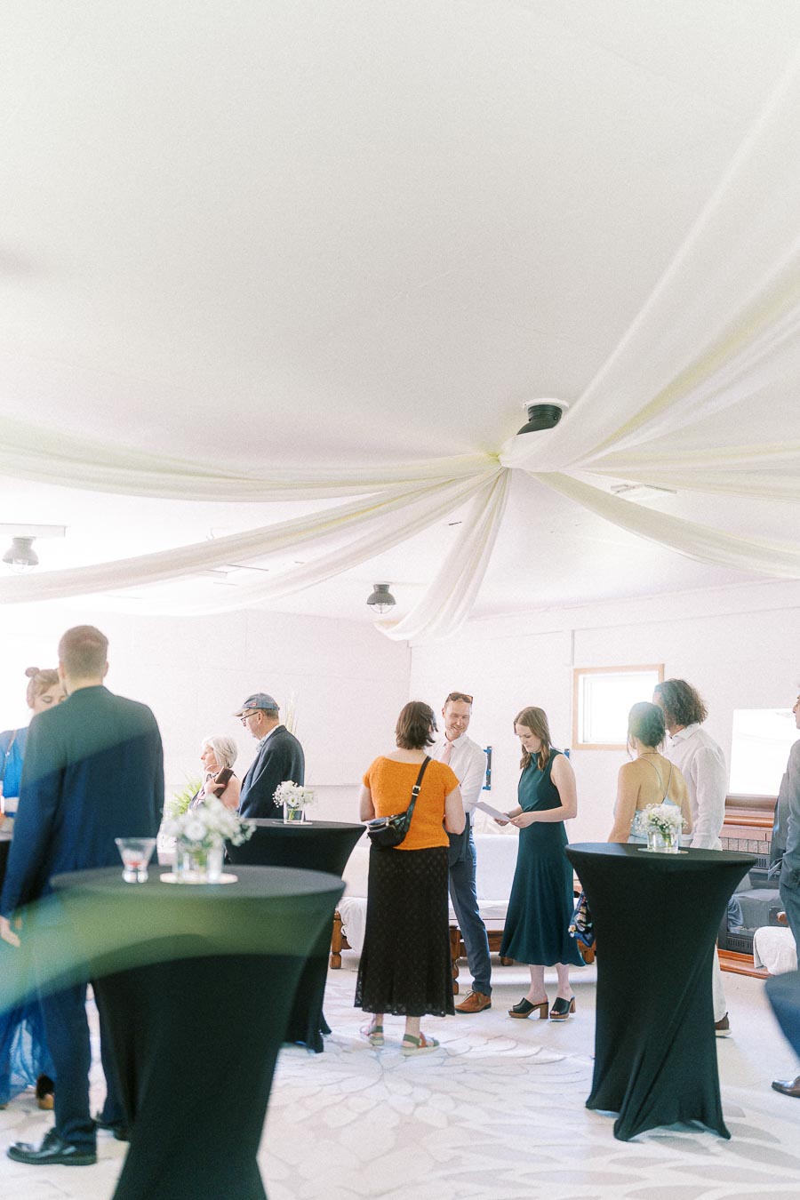 Group of people socializing at an indoor event with elegant white draped ceiling and cocktail tables, creating a casual and sophisticated atmosphere.
