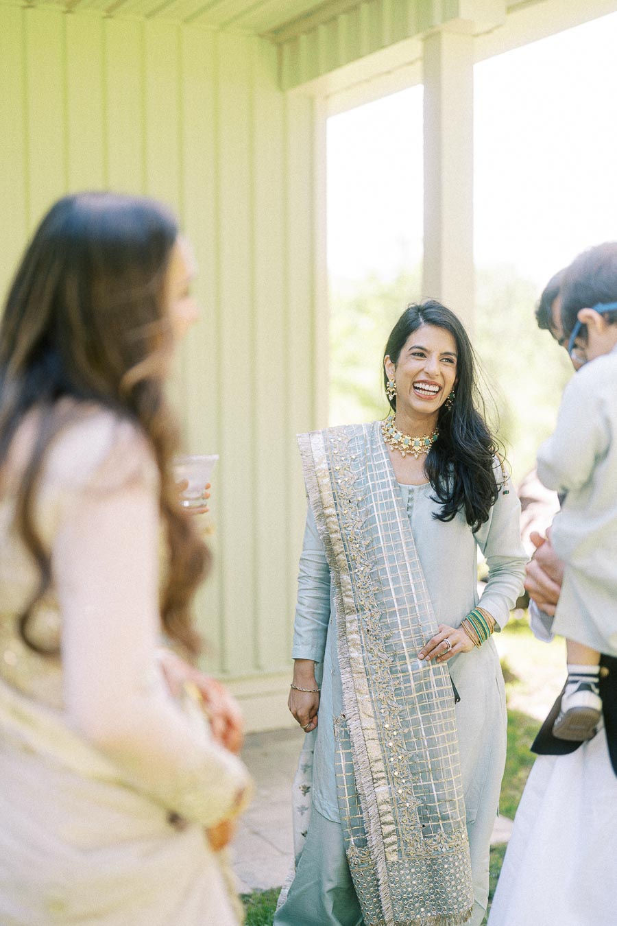 Smiling woman in light blue traditional attire, adorned with a gold and silver-embroidered dupatta, enjoying an outdoor gathering with friends.
