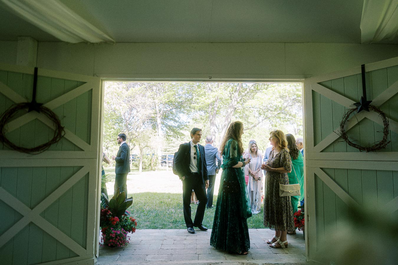 Elegant garden party entrance featuring guests in formal attire under sunny skies, framed by open barn doors adorned with rustic wreaths, suitable for outdoor event promotions.