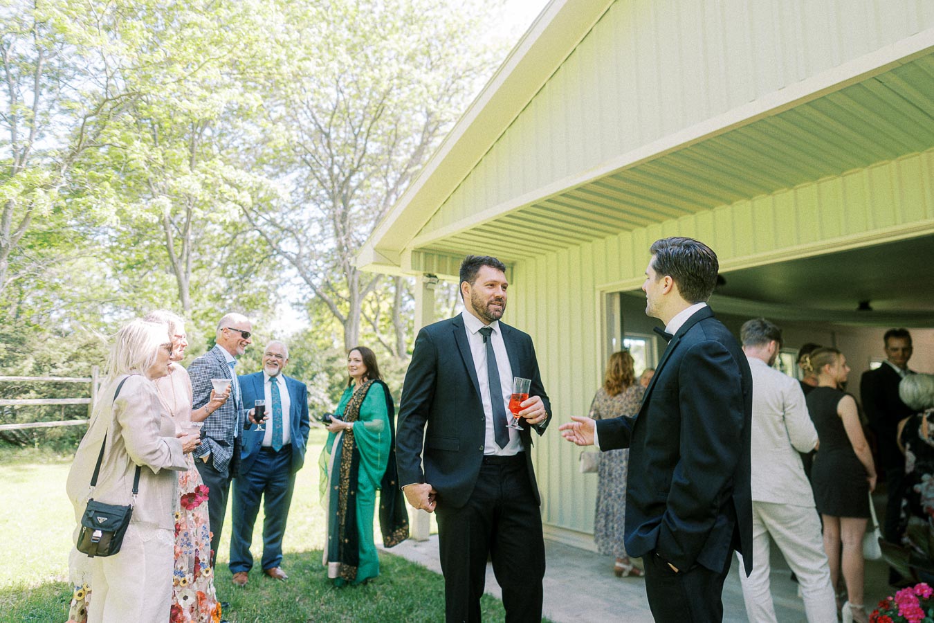 Group of elegantly dressed people socializing outdoors at a formal event, near a light green building, with greenery and trees in the background.