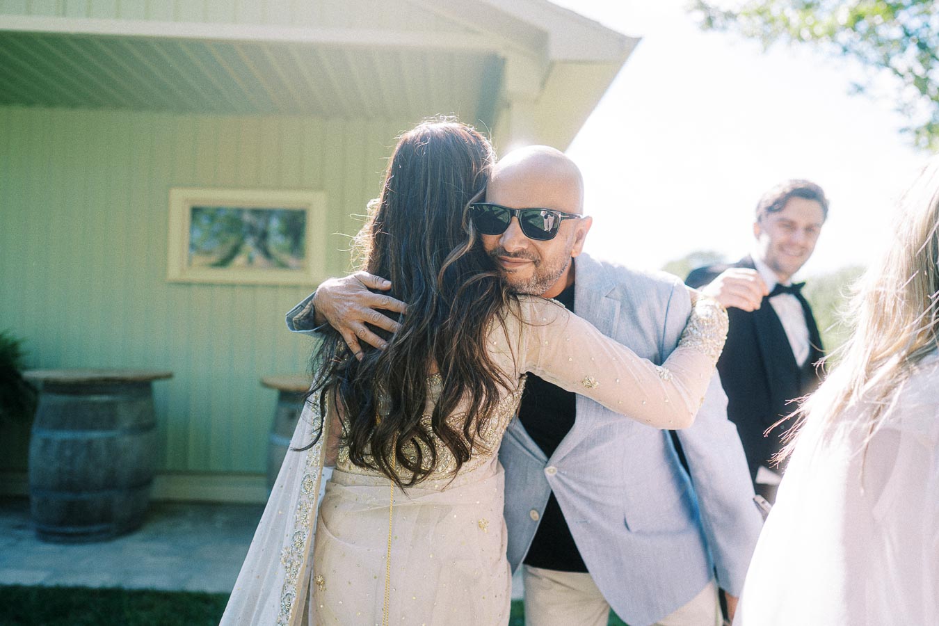 Two people warmly embracing at an outdoor event, with a man in a suit and sunglasses smiling, capturing a joyful moment of celebration and connection.