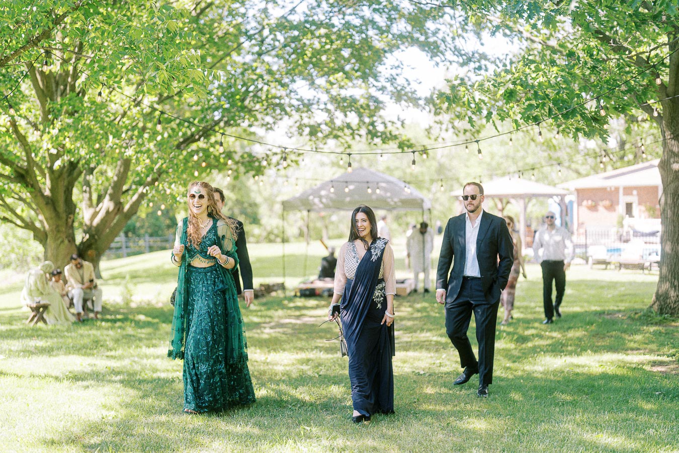 Group of elegantly dressed people walking outdoors at a sunny garden party, featuring lush greenery, party lights, and a festive atmosphere.