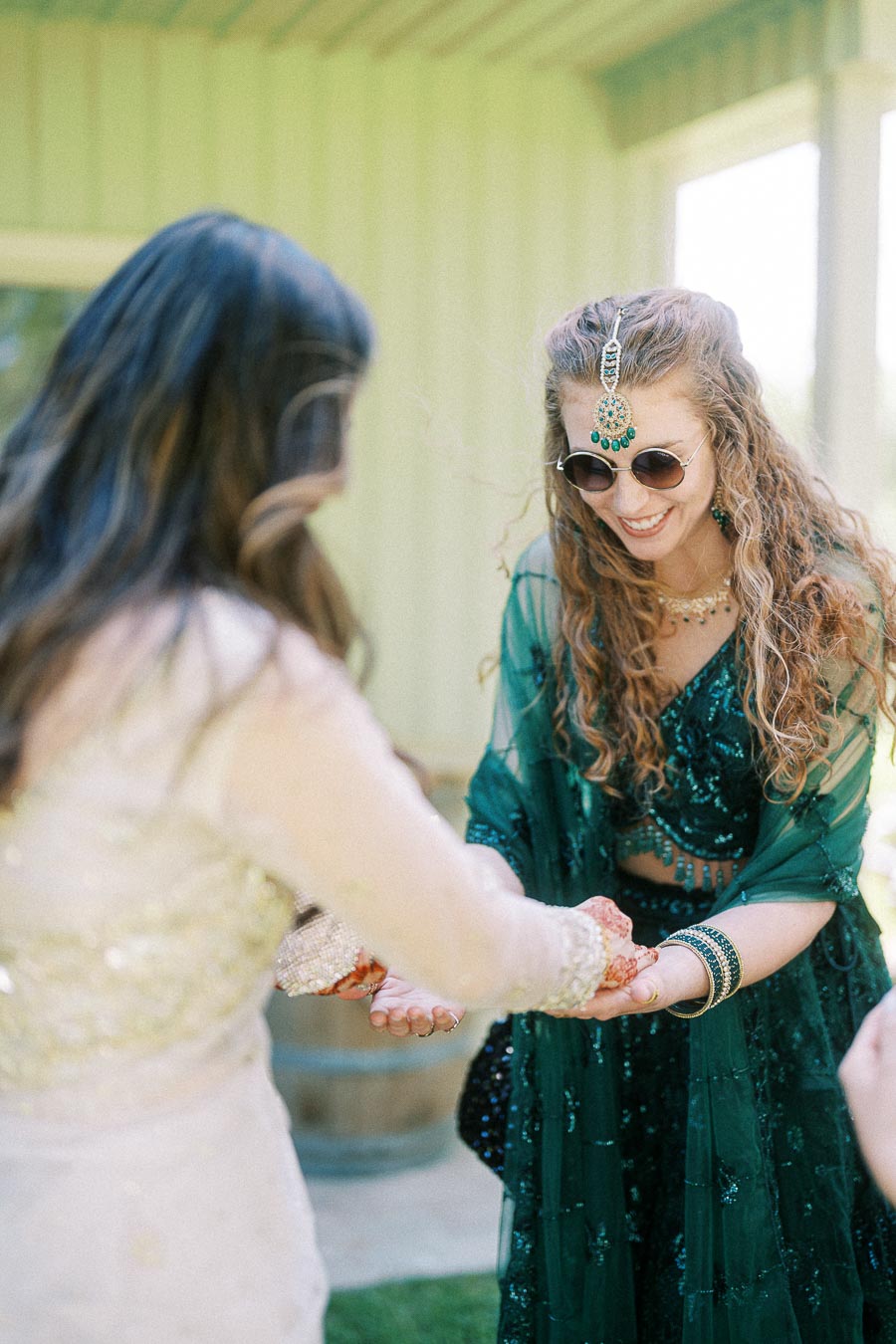 Two women enjoying a festive celebration, one wearing a green embroidered outfit with traditional jewelry and sunglasses, while the other has her hands adorned with henna, capturing a joyful moment outdoors.