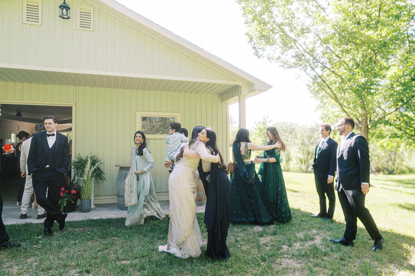 A group of people dressed in formal and traditional attire are engaged in a joyful gathering outdoors near a light-colored house, with some embracing and others conversing, surrounded by greenery on a sunny day.