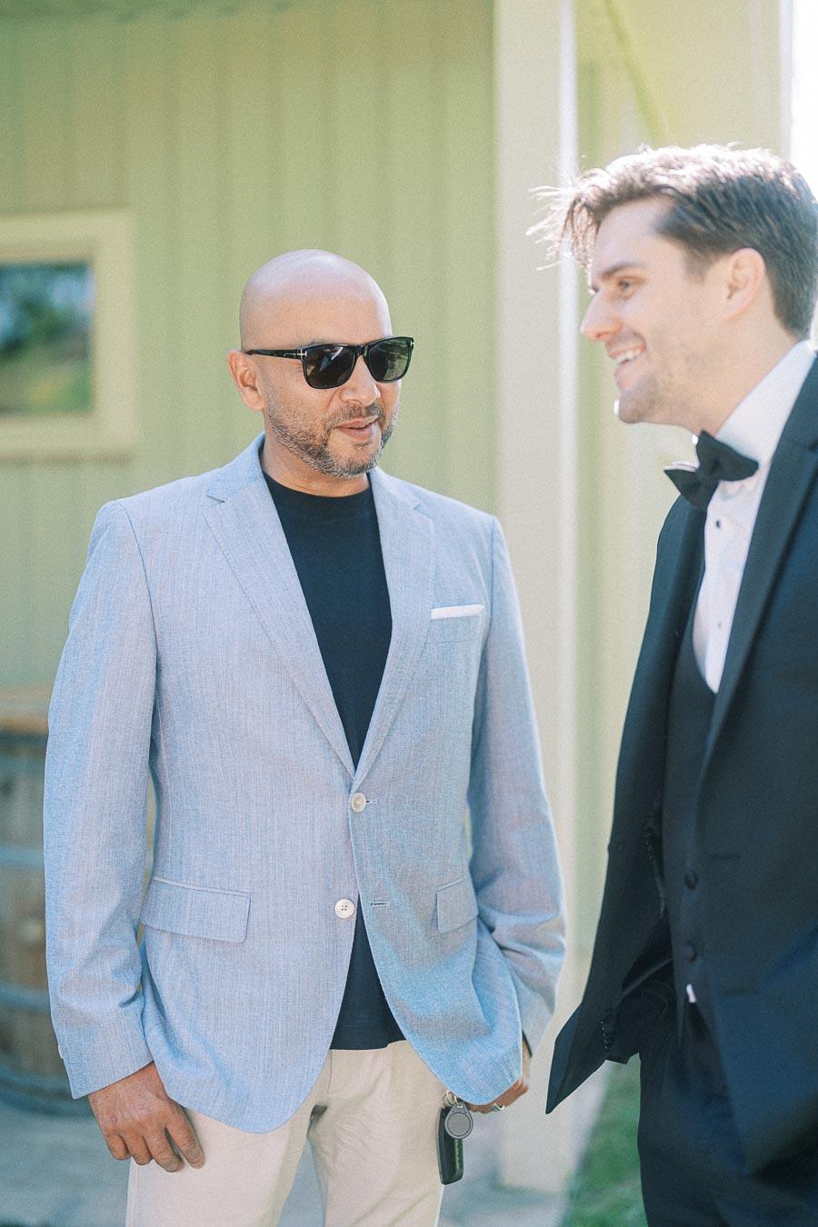 Two men engaged in conversation outdoors, one wearing sunglasses and a light grey blazer, the other in a formal black suit with a bow tie, standing in front of a building with light green siding.