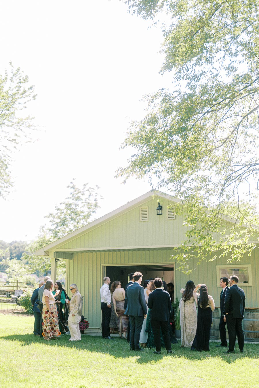 Outdoor gathering at a light green barn with people in formal attire on a sunny day, surrounded by lush greenery.