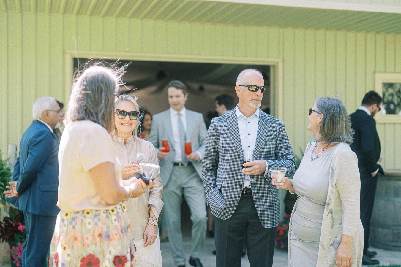 Group of elegantly dressed people socializing outdoors at a formal gathering, engaging in conversation and holding drinks, with men in suits and women in stylish outfits, highlighting a sophisticated event atmosphere.
