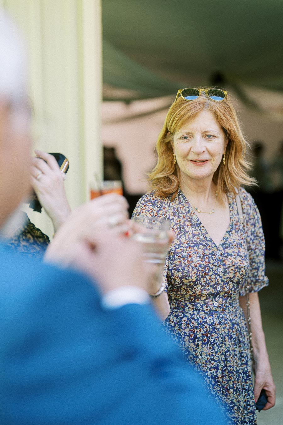 Woman in floral dress socializing at outdoor event, holding a drink and engaging with others, wearing sunglasses on head.