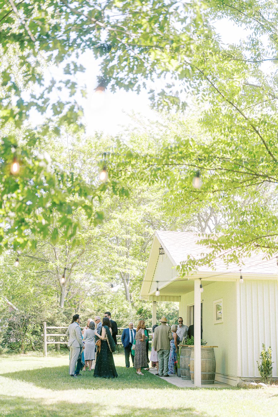 Group of people gathered outside a charming wooden building in a lush, green garden setting, surrounded by trees and string lights on a sunny day.