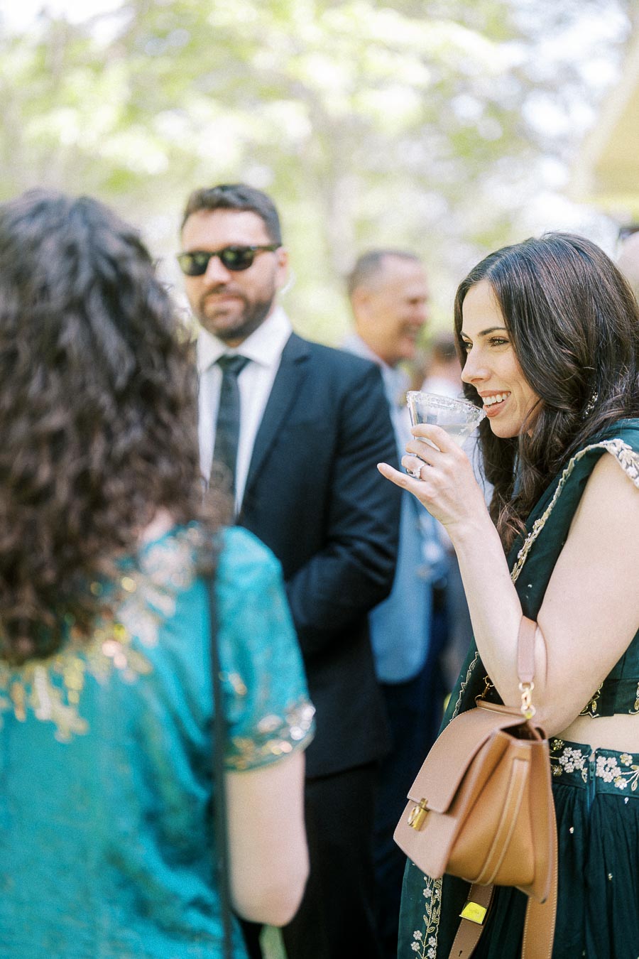 Elegant outdoor gathering with people socializing, featuring a woman in a green dress smiling while holding a drink, surrounded by well-dressed individuals in a sunny, lush setting.